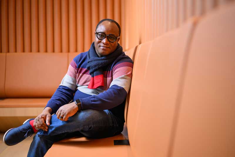 Ericmoore Jossou sits with legs crossed on an orange couch, with orange walls in background