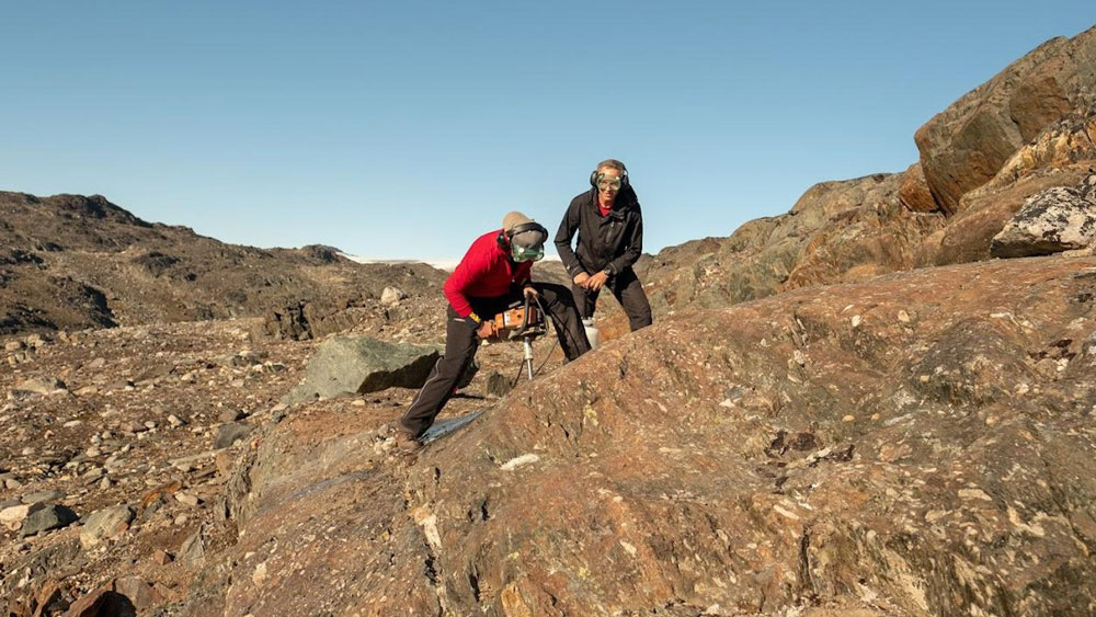 On a vast rocky landscape, Claire Nichols drills into brown rock as Ben Weiss smiles. Both wear protective goggles.