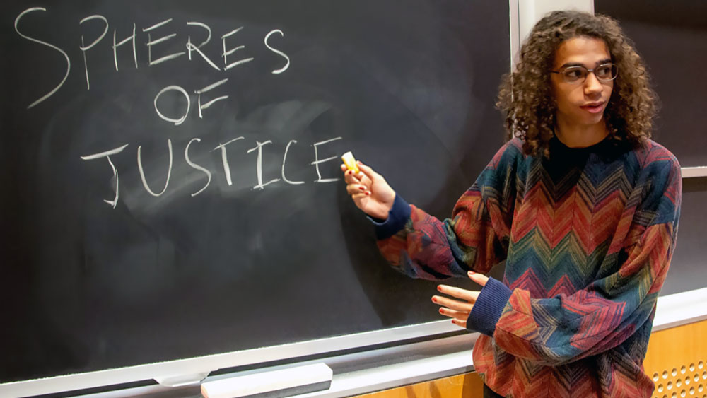 A photograph of an MIT student standing at the chalk board in the Spheres of Justice Class