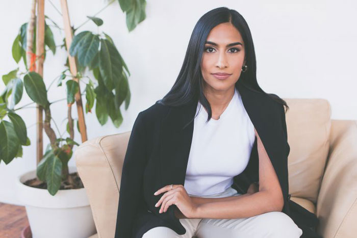 Photo of Kirin Sinha indoors sitting on chair with plant in background