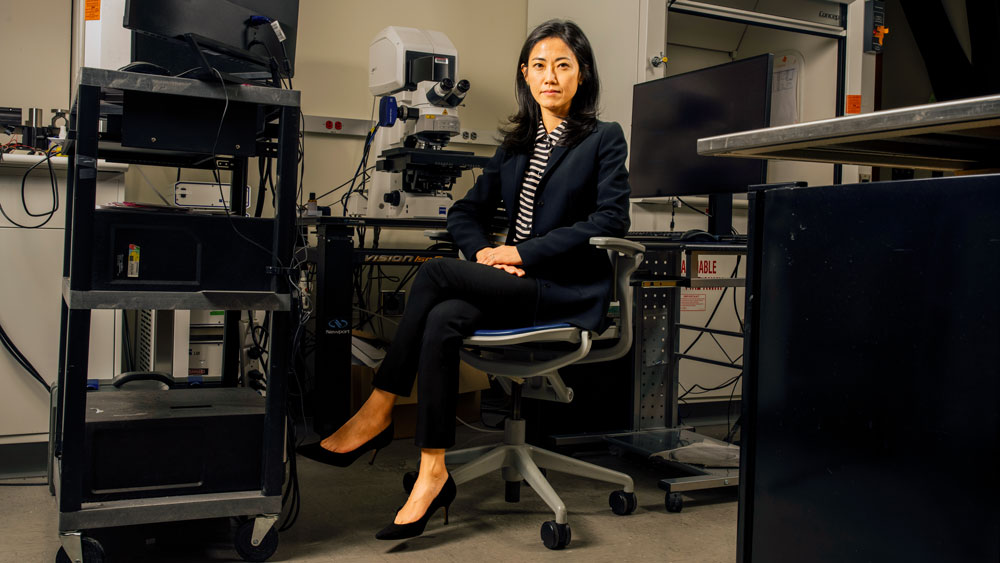 Gloria Choi sits on a chair surrounded by grey and black equipment in a lab, including a microscope, computer monitors, and rolling carts with devices on them. 