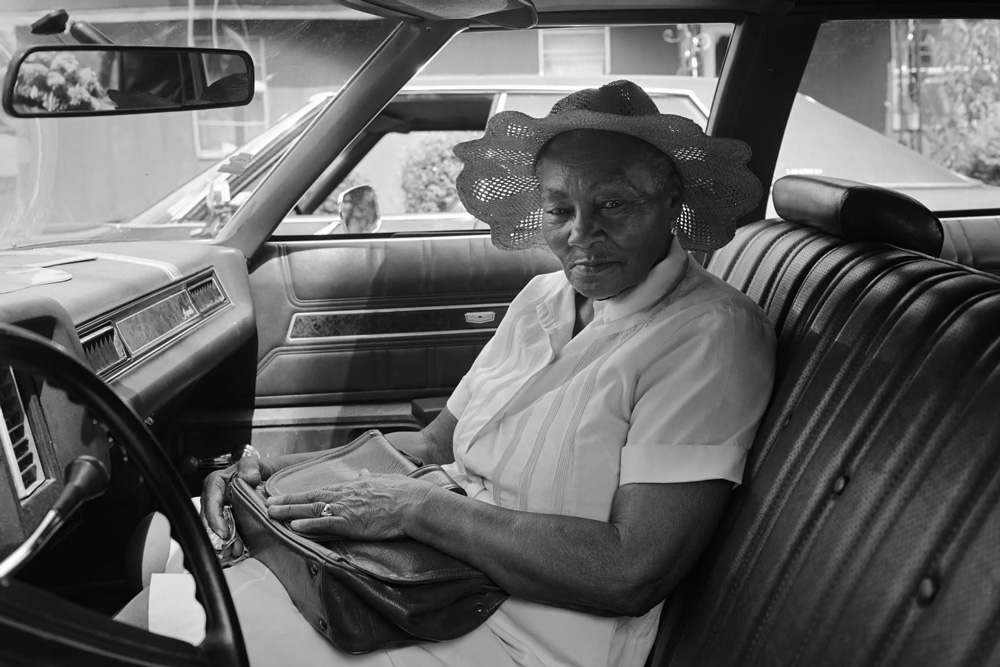 1984 black and-white photo shows an older woman sitting in a car, purse in her hands, wearing a hat and a light-colored uniform. 