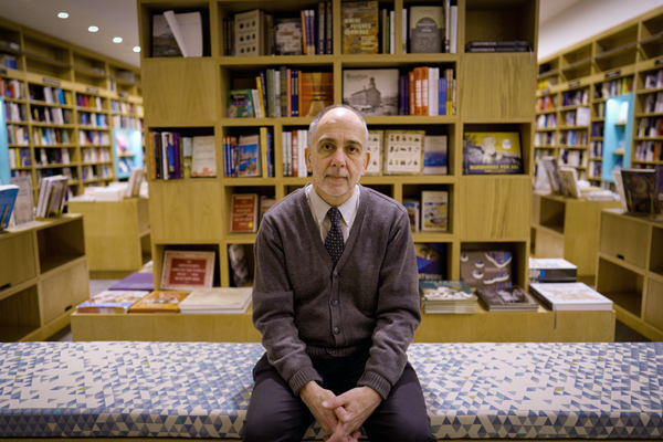 Barry Duncan sits on a bench inside MIT Press bookstore, with shelves of colorful books surrounding him.