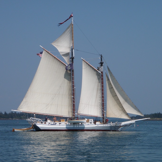 A two masted sailboat, fore-and-aft rigged, with two large gaff rigged sails.  The aft mast is taller and has a triangular sail above the gaff riged one.  The boat has two jibs.  A smaller boat is attached to the stern, and another smaller boat hangs by the rail at the center of the ship.  A pennant flies from the top of the aft mast, and an American flag flies from the aft edge of the end of the aft gaff.
