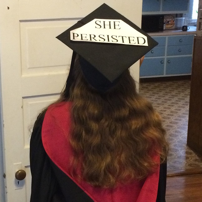 A person wearing graduation robes faces away from the camera.  The robes are black, the master's hood is lined in red.  The person's hair falls to the middle of their back.  The mortarboard is decorated with the words 'SHE PERSISTED'.