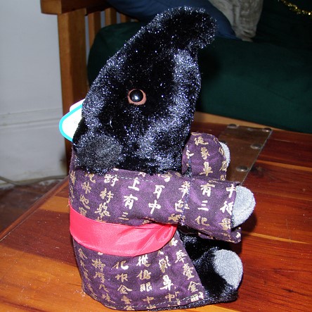 A small stuffed yumekui wearing a purple kimono with fuschia sash.  The kimono is decorated with gold characters.