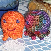 Two small crocheted octopi.  The one on the left is orange with blue eyes and a big grin.  The one on the right shades from pink on the upper left to purple on the lower right and has widely spaced blue eys and pursed red lips.