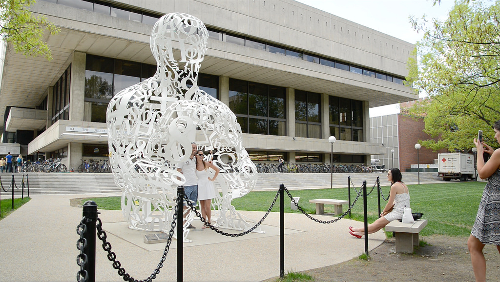 People pose for photos in front of “Alchemist.” The white sculpture is in the shape of a person's head and torso and is made up of numbers and symbols.