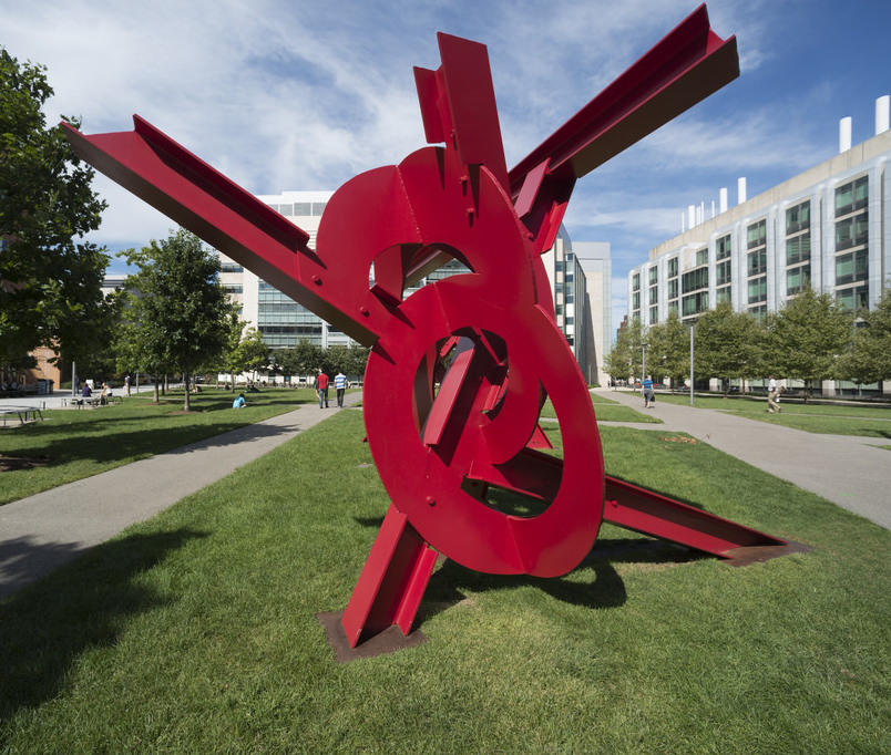 A large, red sculpture sits on the grass. MIT buildings and people walking are seen in the background.