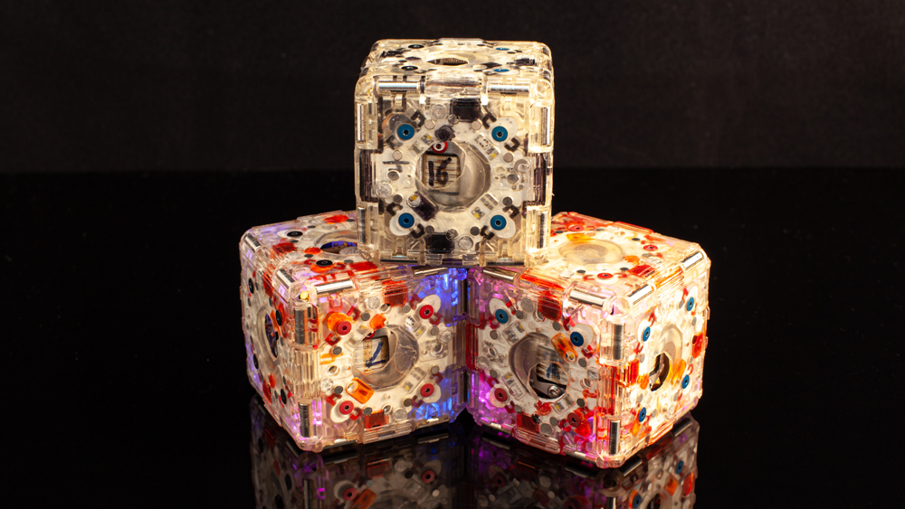 3 m-block robots with different colored components encased in clear plastic and stacked in a pyramid on a black surface