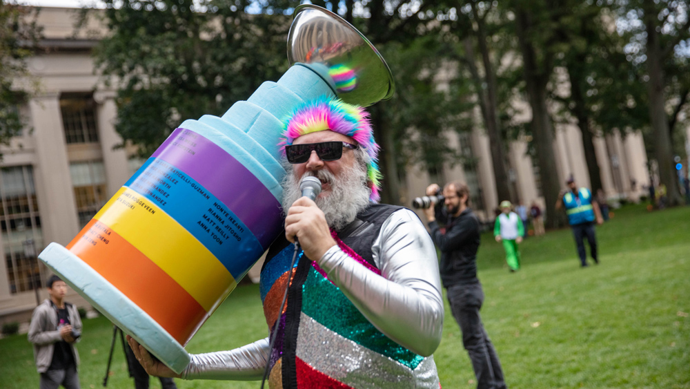 David Wallace wearing sunglasses, and a rainbow wig and vest and carrying a large striped trophy