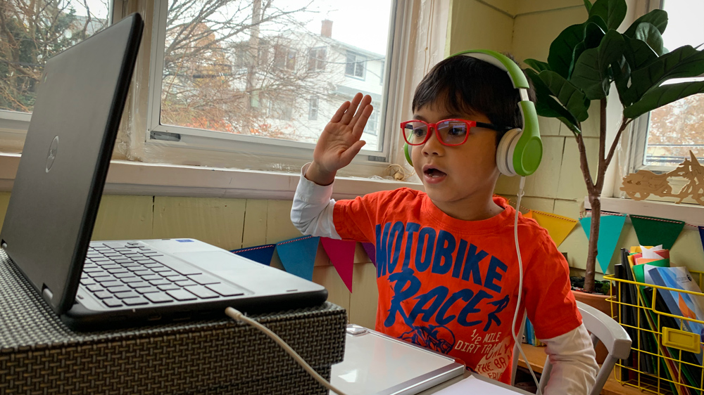 boy in orange shirt, green headphones and glasses raising his hand in front of a computer during a remote learning session