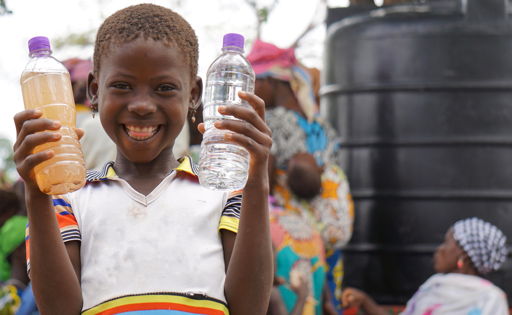 a small, smiling Ghanaian child holds two water bottles; one full of clean water and one full with dirty brown water