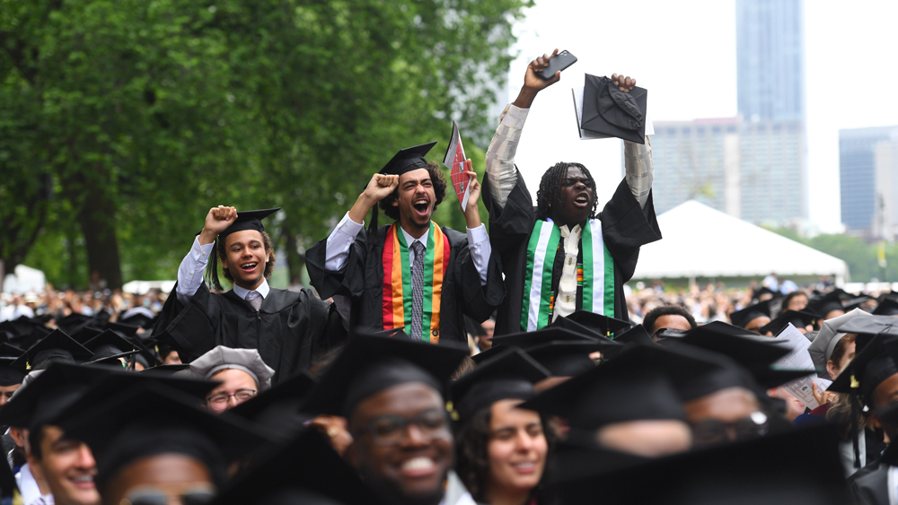 three MIT graduates standing above the crowd of graduates in the audience with their hands in the air