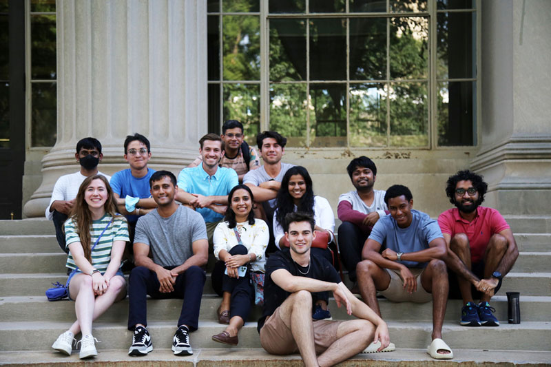13 grad students smile and sit on the steps of Building 10 on Killian Court