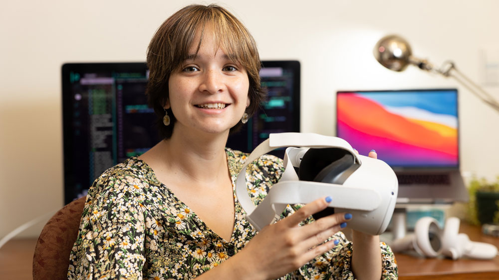 Fernanda De La Torre sits at a desk holding an Oculus Virtual Reality headset, with computers in background.