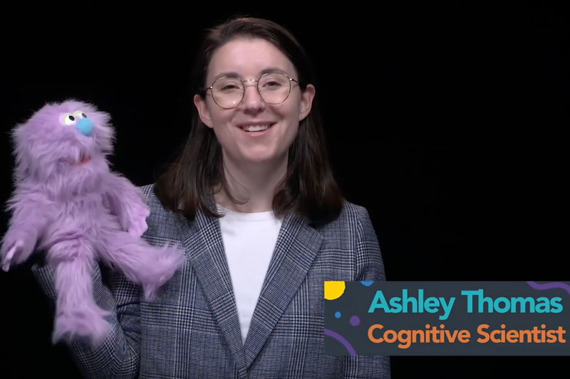 Ashley Thomas holds a furry light purple puppet with a teal nose. The text, "Ashley Thomas, Cognitive Scientist," is on the bottom right.