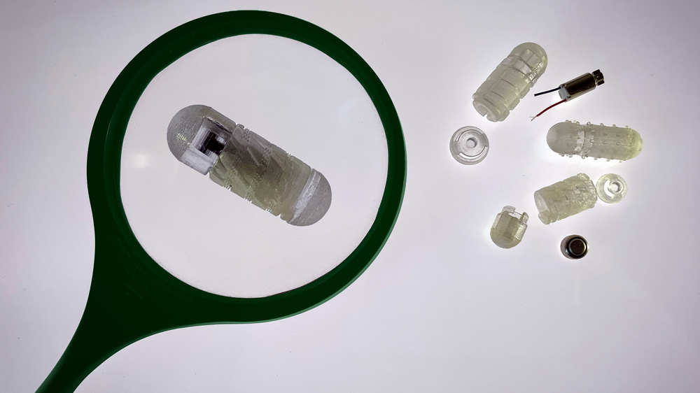 Pieces of the drug capsule prototype are on a grey background. Some pieces are pill-shaped and made of translucent material. Some are threaded like a screw. On the left, a green magnifying glass enlarges one pill-shaped device.