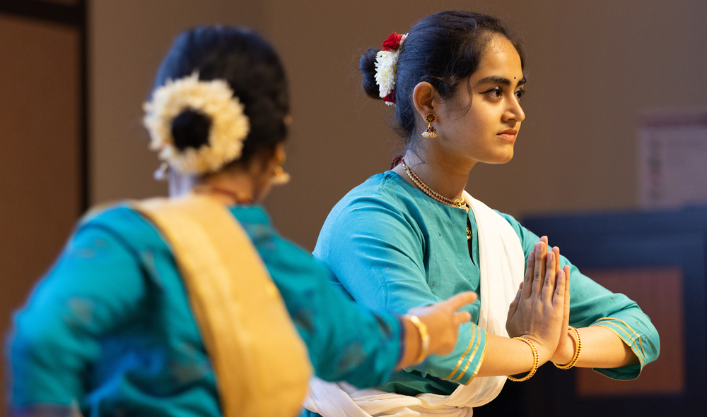 2 dancers with teal garments, gold jewelry and eye makeup, one holding her hands pressed together.