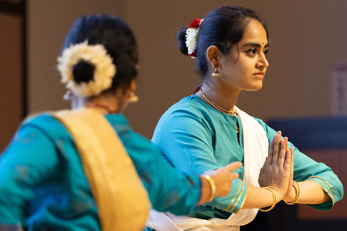 2 dancers with teal garments, gold jewelry and eye makeup, one holding her hands pressed together.