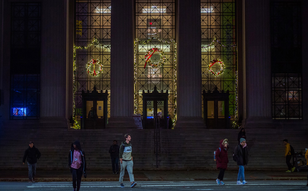 Tall windows at MIT at night decorated with wreaths and garlands while people walk in the foreground