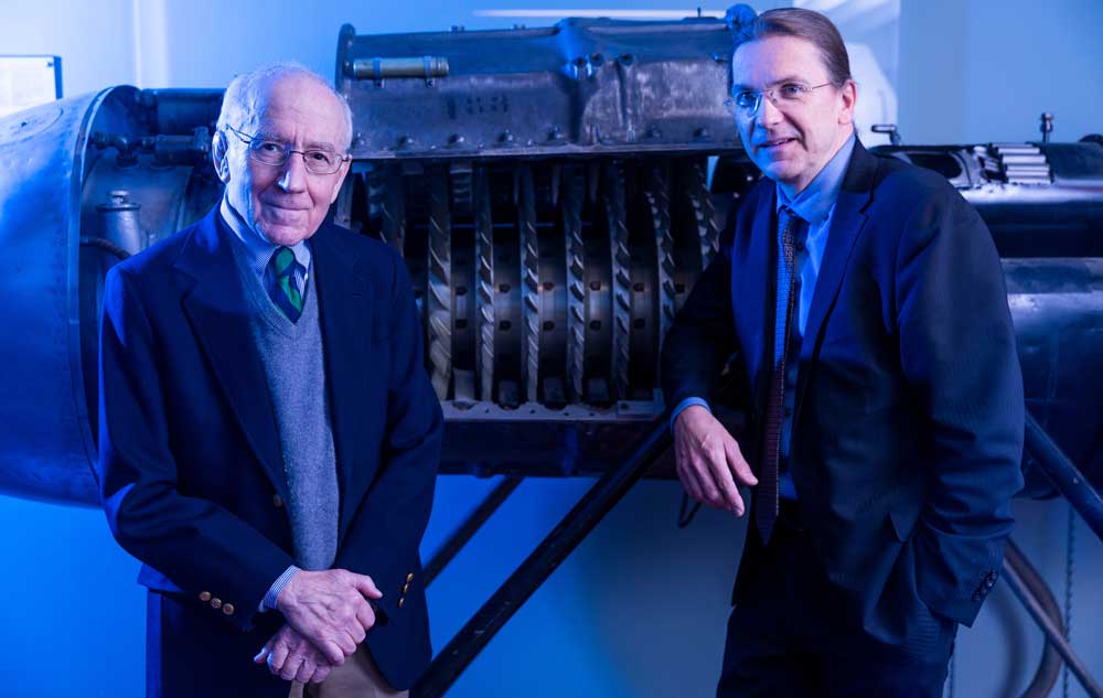 Zoltán Spakovszky and Edward Greitzer lean against large turbine engine machinery on display, with blue lighting