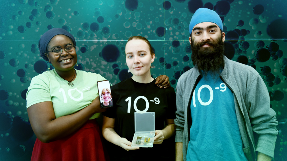 (Left to right:) Senior Stephanie Khaguli, junior Vlada Petrusenko, sophomore Anhad Sawhney, and sophomore Matthew Taylor (not photographed but appearing on Khaguli's phone). and Photoshopped teal molecules surround them. Khaguli holds a phone with their group member smiling. Petrusenko holds a device that looks like 3 computer chips inside a plastic case.