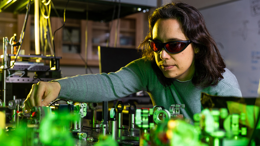 Schlau-Cohen, in a laser lab wearing special glasses, tweaks a device on a table. The room is dark, and the tabletop has green light and many shiny laser parts.