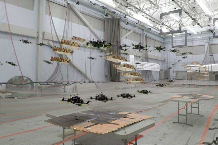 Combined video stills show the pathways of four flying drones in a large auditorium. The four drones avoid two flying obstacles, which are drones with a camouflage box around then.