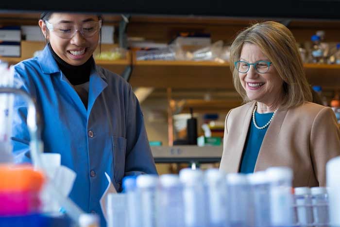 Kornbluth, on right, and Yang smile while inside a busy lab. Blurry equipment is in the foreground.