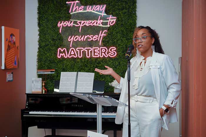 Lauryn McNair speaks on stage, and in background is a neon sign saying, "How you speak to yourself matters."