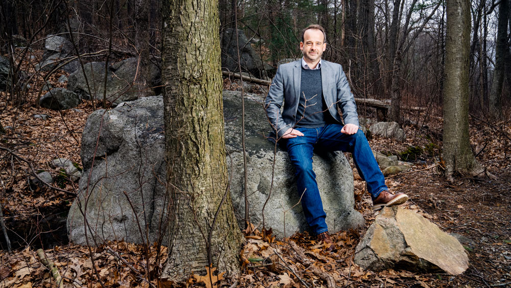 Fournier sits on a large grey rock in the woods. The textures of the trees, rocks, and dead leaves contrasts with Fournier’s blue jeans.
