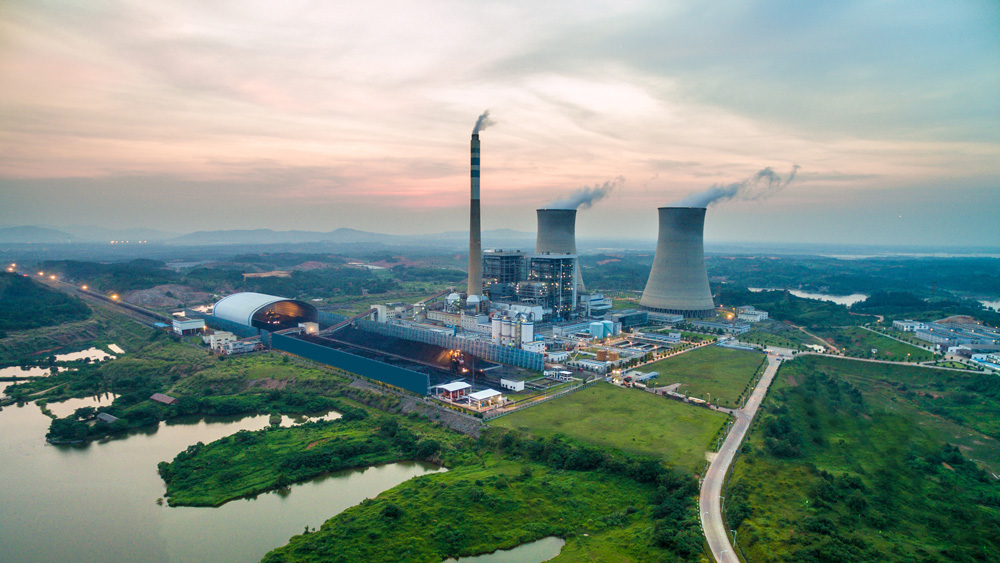 Aerial view shows a nuclear power plant, surrounded by lush green areas and water, with mountains in distance.