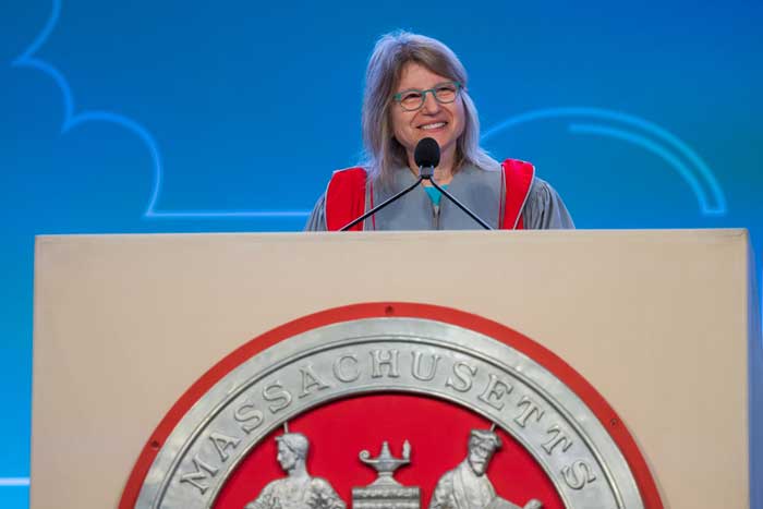 President Kornbluth, wearing grey and red academic regalia, speaks at a podium with a large MIT seal on the front.