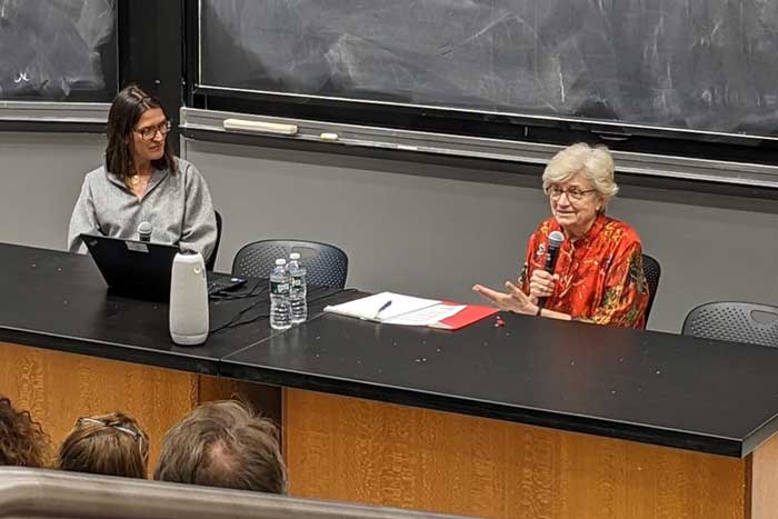 Kate Zernike and Nancy Hopkins sit at a table addressing an audience in a classroom. A blackboard appears behind them.