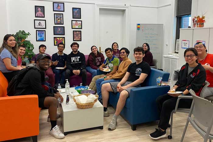 About 14 MIT community members relaxing inside eating lunch and sitting on couches, and smiling for the photo