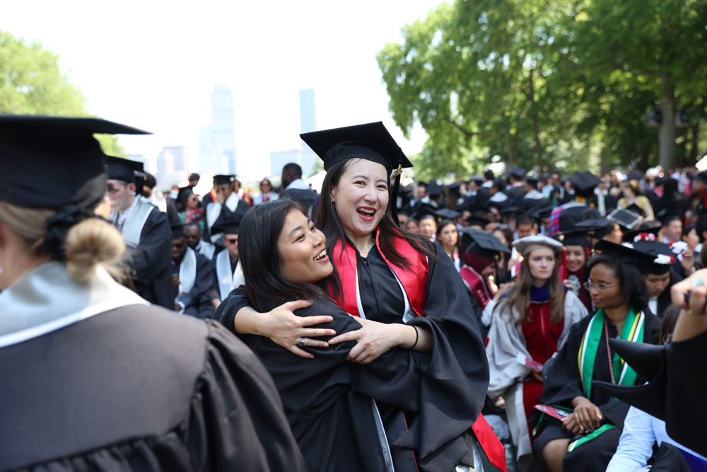 2 recent graduates hug with joy while surrounded by graduates. The Boston skyline is in the background.