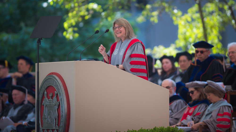 Kornbluth at the podium speaking to the crowd, with green foliage and faculty in background