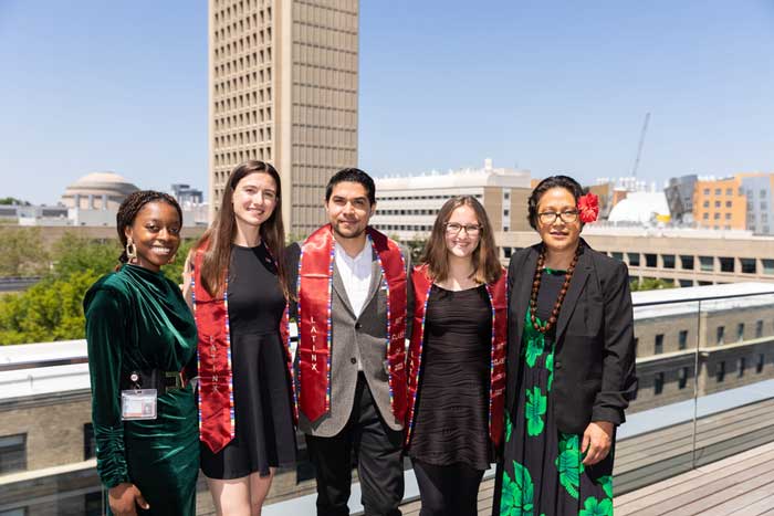 Five individuals pose together on a balcony with MIT campus buildings in the background. Three wear red stoles around their shoulders.