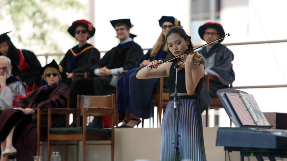 Shelley Choi playing the violin on stage while MIT faculty members watch behind her