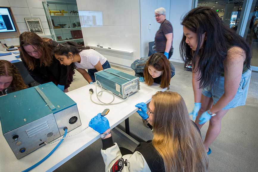 6 students surround a vintage cardiac arrhythmia detector, examining it closely.