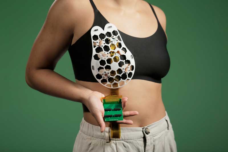 Photo shows a woman wearing the breast cancer-detecting prototype device over black tank top on a green background. In her hands she shows the green circuit board. 
