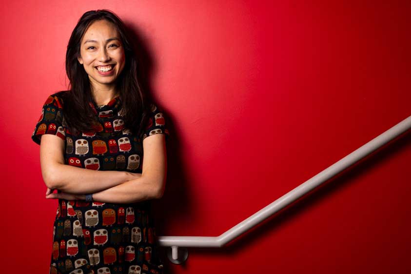 Lisa Ho smiles while leaning against a red stairwell wall. A grey handrail diagonally goes up.