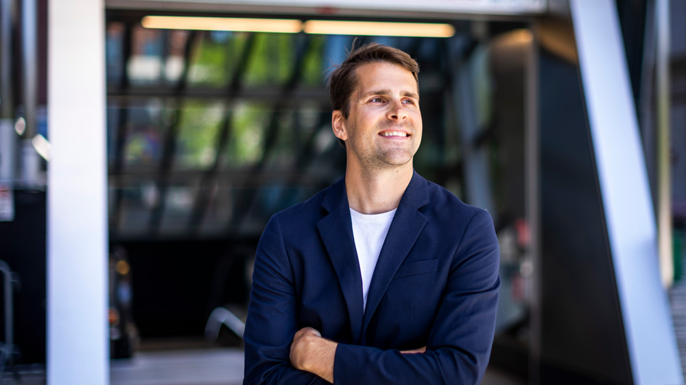 Caros smiles and stands near the exit of the Kendall/MIT station near campus. You can see a bit of the escalator and skylights in background.