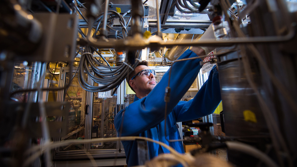 Jamison Watson, wearing safety glasses, adjusts the trickle-bed reactor, comprising a variety of pipes and wires in the foreground of the image.