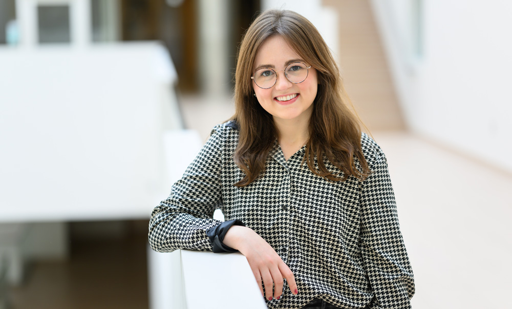 Portrait photo of Kathrin "Kat" Kajderowicz leaning against a railing, with white walls and stair case in background