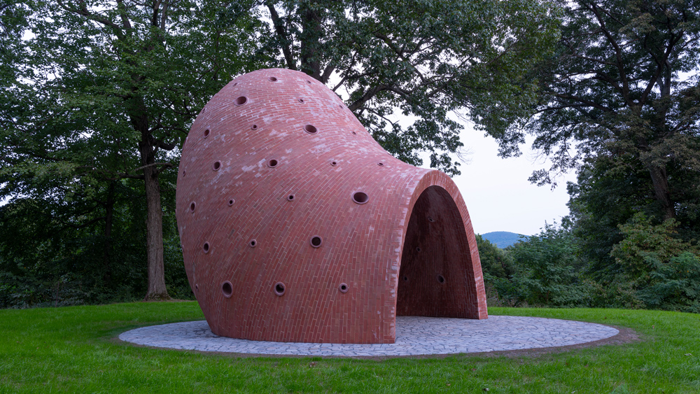 A side view of the red brick sculpture. It has an arched entryway, and then curves upward to an off-center dome ceiling. It has small circular windows scattered throughout.