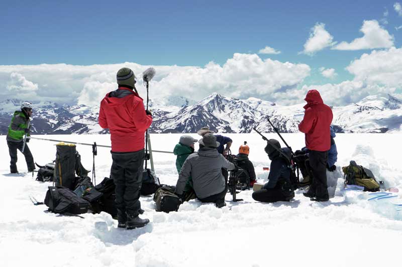 About 9 people pause in a vast snowy, mountainy landscape with camera and sound equipment