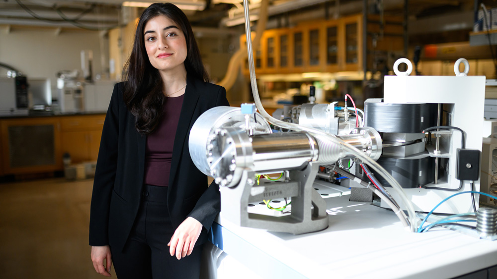 Fatima Husain stands in a lab with arm resting on table. The table has a complex machine with a cylindrical base and tubes and wires connected to the table and ceiling.
