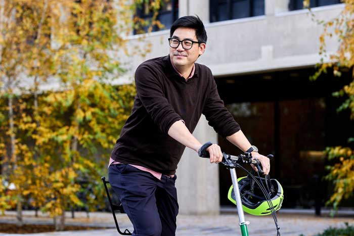 David Hsu rides a bike, which is half-seen, while outside and with yellow foliage in the background